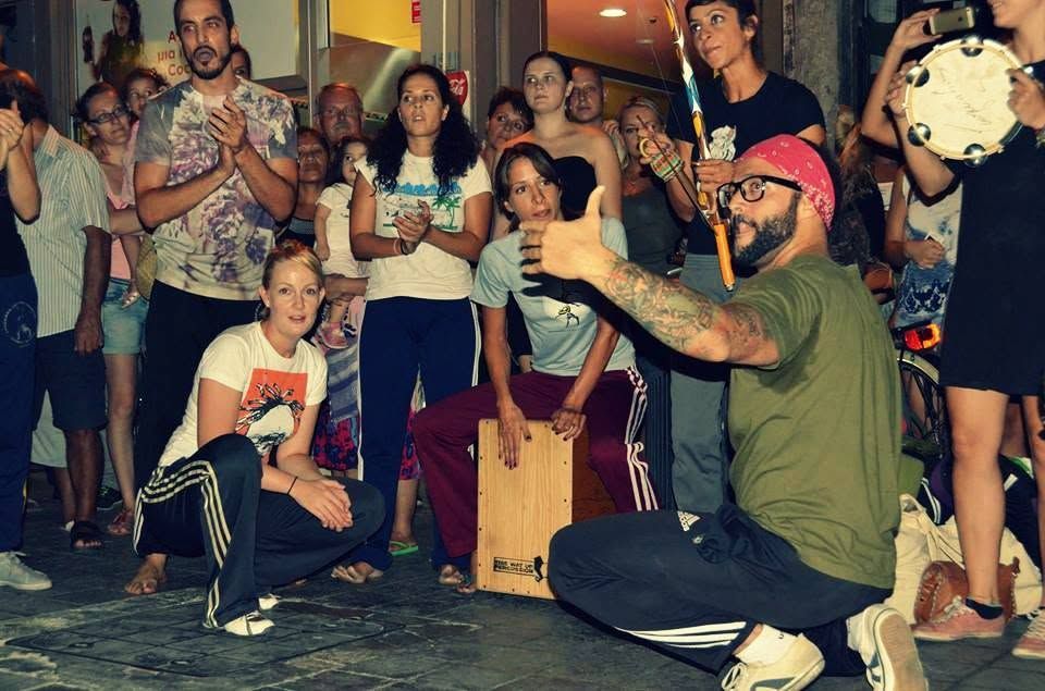 Mestre Casquinha giving a thumbs up at an Apêiara street performance on Ledras Street, with a crowd watching